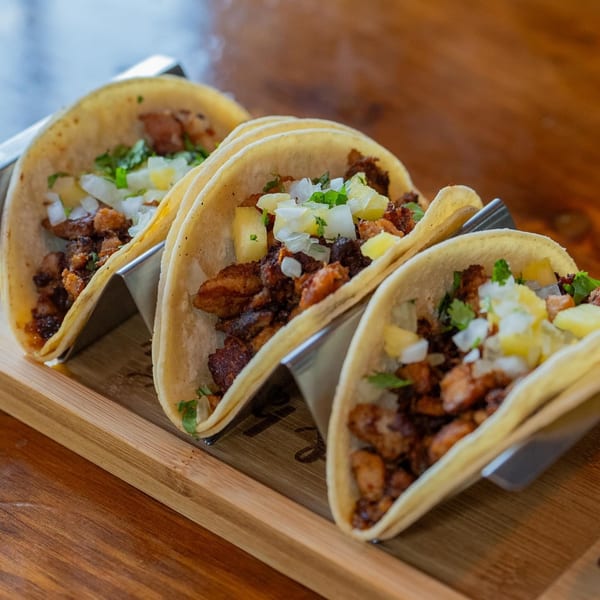 Three al pastor tacos with pineapple, onions, and cilantro, served on a wooden board.