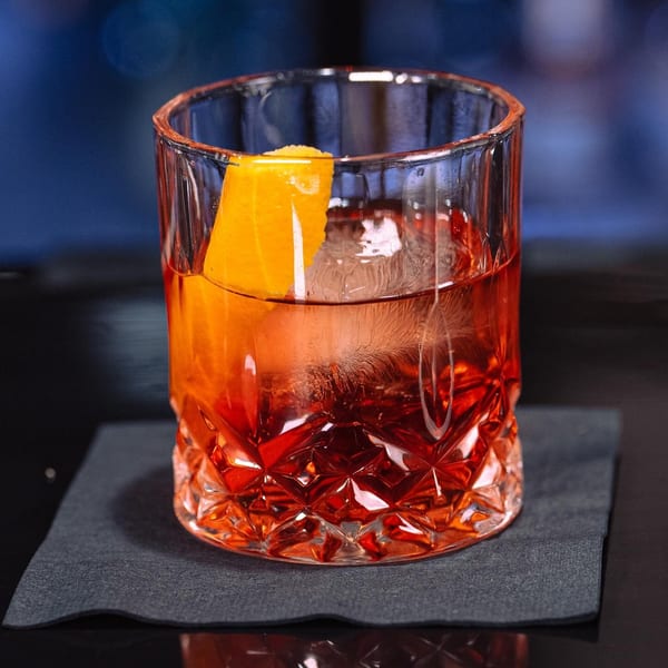 A vibrant red cocktail with a large ice cube and an orange peel garnish in a cut glass.