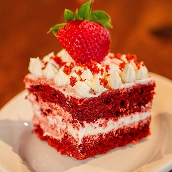 A slice of red velvet cake with cream cheese frosting, topped with a fresh strawberry.