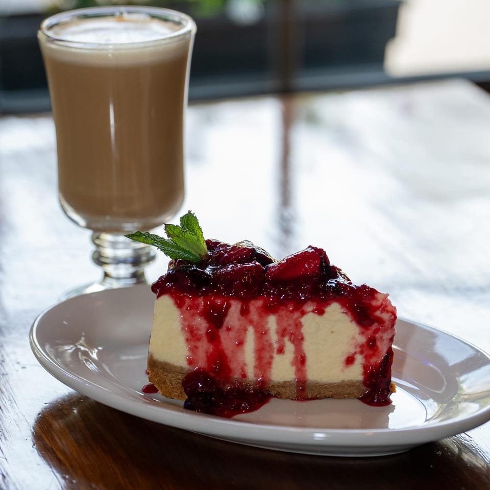 A slice of cheesecake with berry topping and mint, next to a glass of iced coffee.