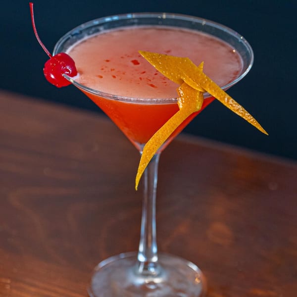 A vibrant red cocktail in a martini glass, garnished with a cherry and orange peel.