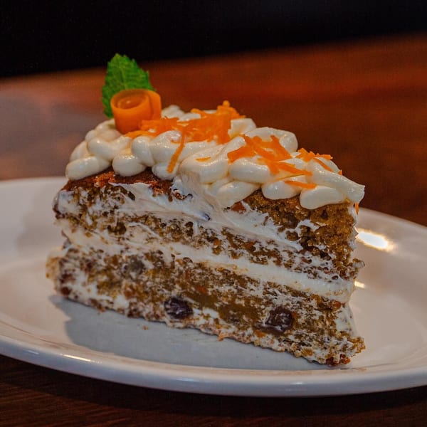 A slice of carrot cake with cream cheese frosting, topped with shredded carrots and a mint leaf.