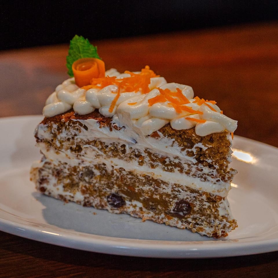A slice of carrot cake with cream cheese frosting, topped with shredded carrots and a mint leaf.