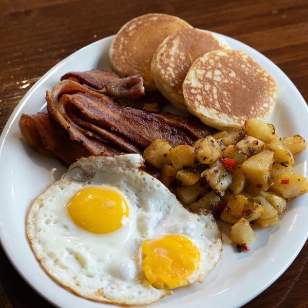 A breakfast plate with pancakes, crispy bacon, two fried eggs, and seasoned home fries.